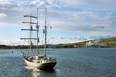 Picture of the tall ship Thalassa anchored in a bay with industrial maltings and a disused distillery in the background