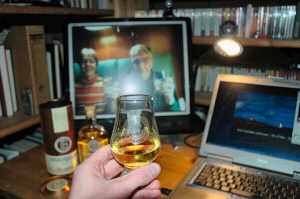 Picture of a dram of whisky being held up in front of a monitor, two people doing the same on the monitor