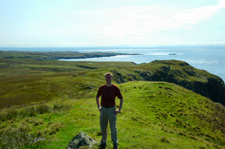 Picture of a man standing on hills near a coast, blue sea under a sunny sky in the background