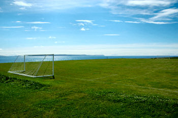 Picture of a football pitch next to a sea loch und a bright blue sky