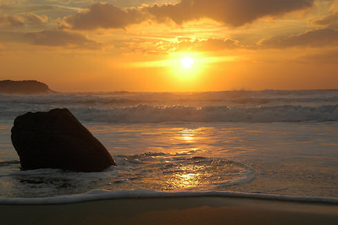 Picture of a sunset over waves crashing at a beach and running up the sand around a rock in the foreground