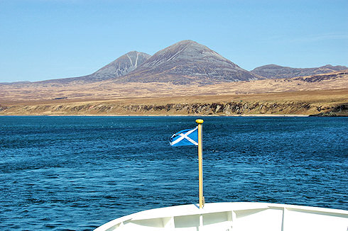 Picture of the Saltire flying on the bow staff of a Calmac ferry with the distinct shape of the Paps of Jura in the background