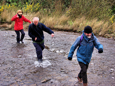 Picture of 3 walkers crossing a wide burn on a windy day