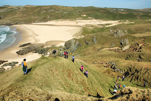 Picture of a long line of walkers descending a hillside towards a sandy beach