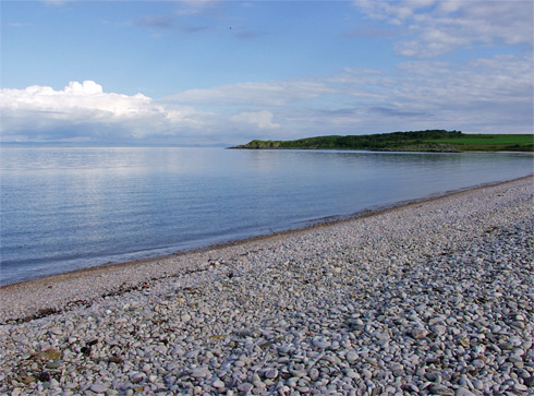 Picture of a bay on a calm day, a pebble beach with a green shoreline in the distance